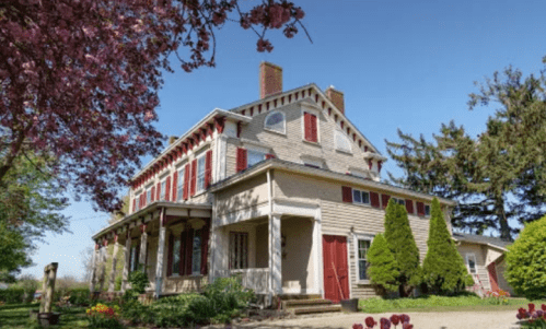 A charming two-story house with red shutters, surrounded by blooming trees and a clear blue sky.