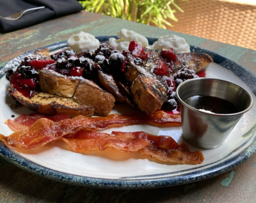 A plate of French toast topped with berries and whipped cream, served with crispy bacon and a small cup of syrup.