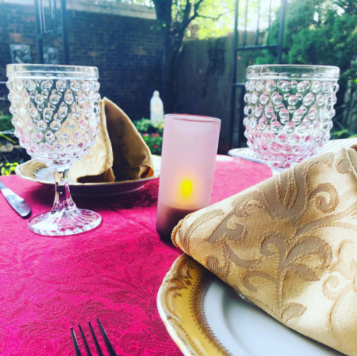 A beautifully set outdoor table with crystal glasses, elegant napkins, and a soft glowing candle on a red tablecloth.