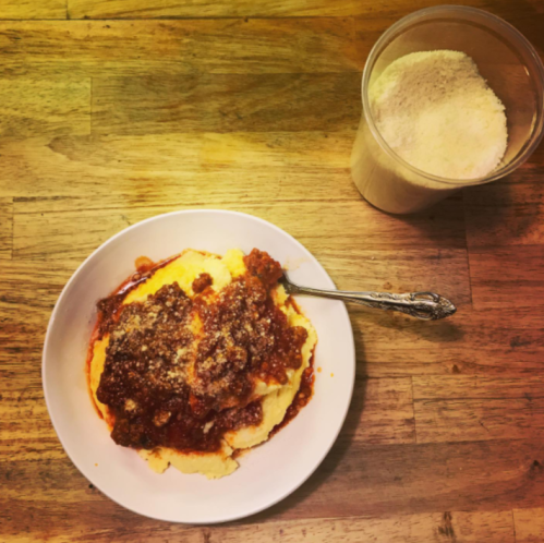 A bowl of creamy polenta topped with sauce and grated cheese, next to a container of grated cheese on a wooden table.