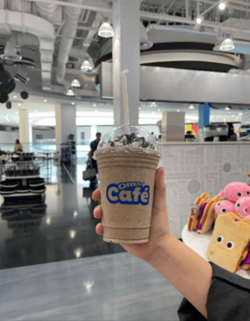 A hand holding an Oreo Café drink in a modern café setting with colorful plush toys in the background.
