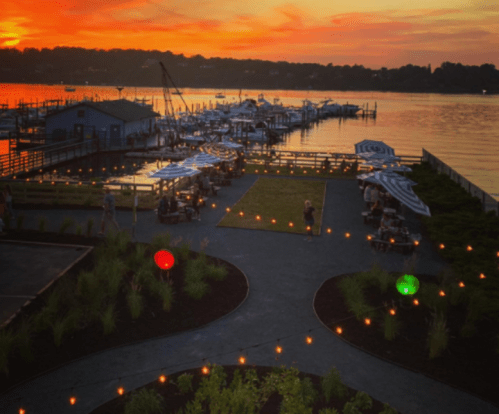 Sunset over a marina with boats, outdoor seating, and colorful lights along a pathway.