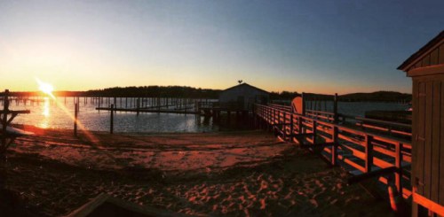 Sunset over a calm waterfront with wooden docks and a sandy beach, casting warm light on the scene.