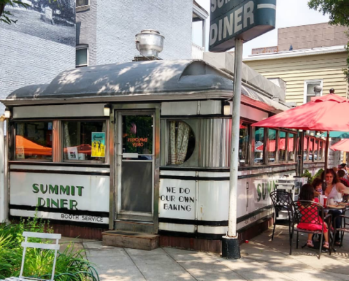 A retro diner with a silver exterior and outdoor seating, featuring umbrellas and patrons enjoying their meals.