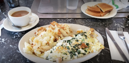 A plate of scrambled eggs with spinach and hash browns, accompanied by a cup of coffee and toast on a table.