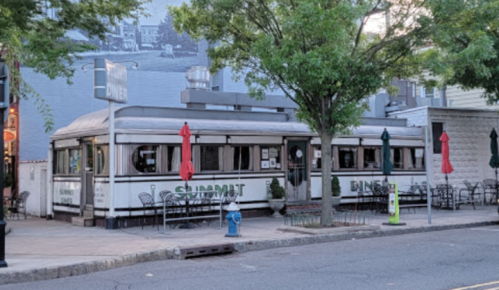 A classic diner with a silver exterior, outdoor seating, and red umbrellas, located on a city street.