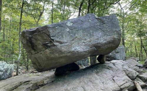 A large, flat rock balanced on smaller stones in a wooded area, surrounded by trees and natural scenery.