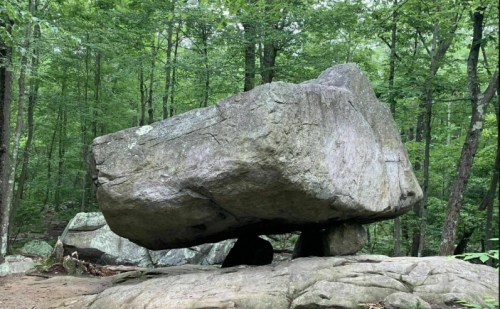 A large, flat rock balanced on a smaller stone, surrounded by lush green trees in a forest setting.