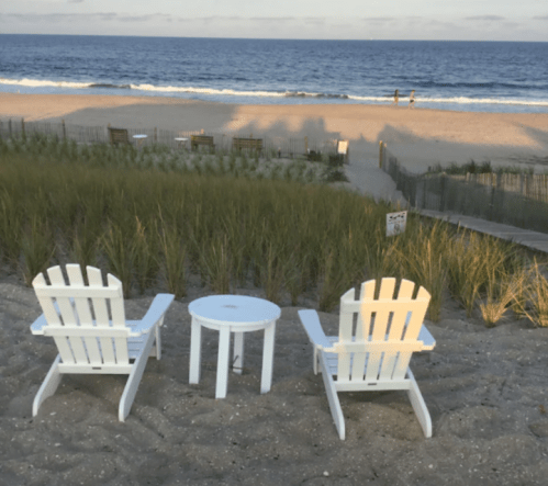 Two white Adirondack chairs and a small table on a sandy beach, with ocean waves and a distant couple walking.