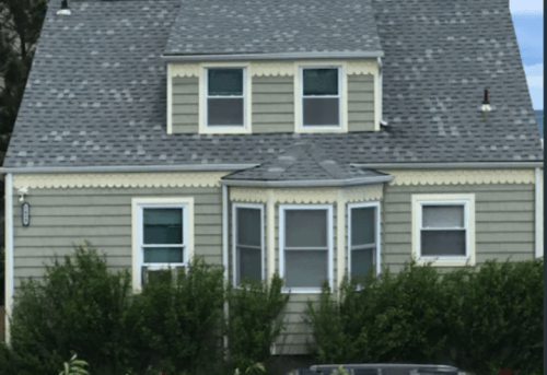 A two-story house with a gray shingle roof and green siding, featuring a bay window and surrounded by shrubs.