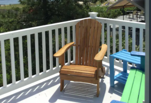 A wooden Adirondack chair on a deck, surrounded by greenery and colorful outdoor furniture.