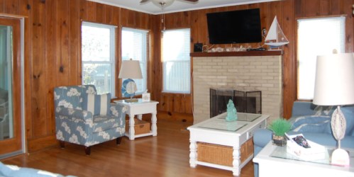 Cozy living room with wooden walls, a fireplace, blue furniture, and large windows letting in natural light.