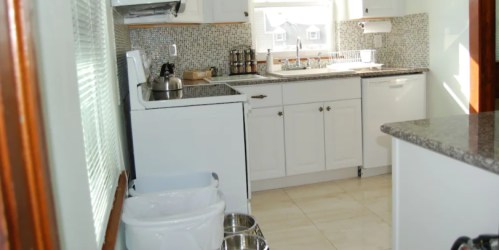 Bright kitchen with white cabinets, a sink, stove, and granite countertops, featuring a window and pet bowls on the floor.