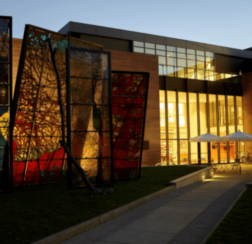 A modern building with large stained glass panels, illuminated at dusk, surrounded by greenery and outdoor seating.