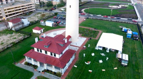 Aerial view of a lighthouse beside a white tent and landscaped lawn, with a parking area in the background.