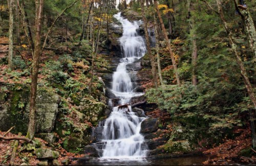 A serene waterfall cascades down rocky steps, surrounded by lush green trees and autumn foliage.
