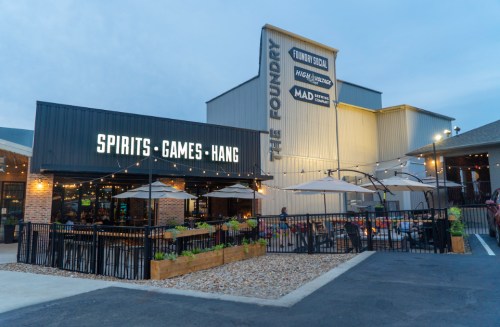 Exterior view of The Foundry, featuring a patio with umbrellas, string lights, and signage for spirits and games.