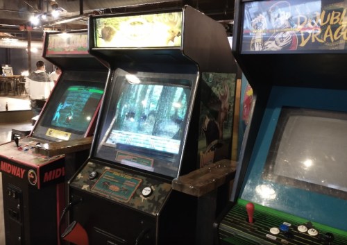 Three vintage arcade machines in a dimly lit gaming area, featuring colorful screens and classic game titles.