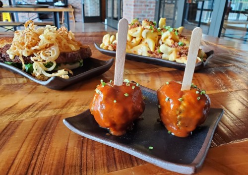 Two meatballs on skewers with sauce, garnished with chives, alongside a plate of crispy onions and another dish of potatoes.