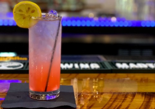 A refreshing cocktail with a lemon slice, served in a tall glass on a bar counter with a colorful background.