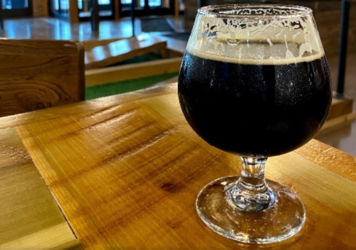 A dark beer in a glass sits on a wooden table, with a frothy head and a blurred background of a bar.
