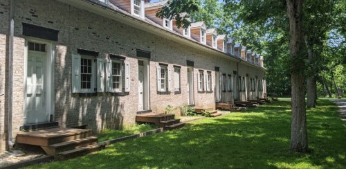 A row of historic brick buildings with white shutters and steps, surrounded by green grass and trees.