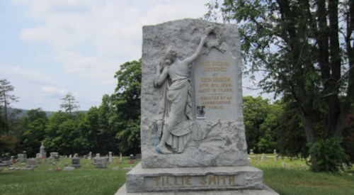 A gravestone featuring a carved figure and the name "Tillie Smith," set in a cemetery with trees in the background.