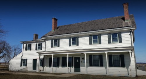 A two-story white house with green shutters and a gray roof, featuring multiple chimneys and a porch.