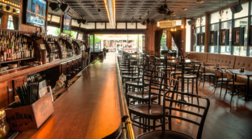 A spacious bar interior with wooden furnishings, empty tables, and a long counter, featuring shelves of liquor and large windows.