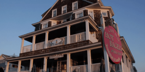 A multi-story building with a sign for "Cape May Hot Bagels" and a porch, set against a clear sky.