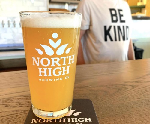 A glass of beer with "North High Brewing" logo on a wooden table, with a person in a "Be Kind" shirt in the background.
