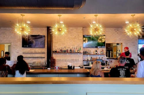A modern bar with stylish lighting, patrons at the counter, and a bartender preparing drinks in a lively atmosphere.