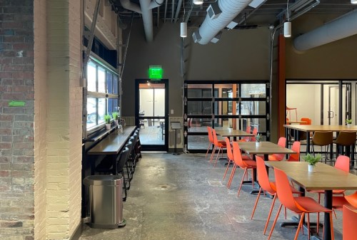 Modern office break room with orange chairs, wooden tables, and large windows, featuring an industrial design.