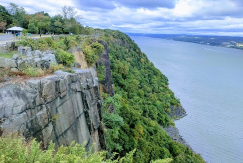 A scenic view of a rocky cliff overlooking a river, surrounded by lush greenery and cloudy skies.