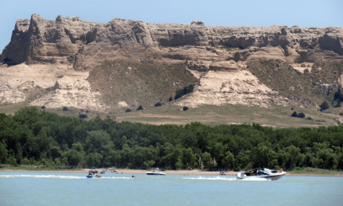 Boats speed across a river with rocky cliffs and lush greenery in the background on a sunny day.
