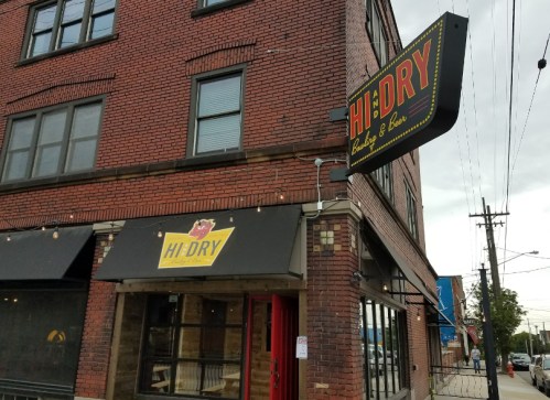 Exterior of Hi-Dry Bar & Bites, featuring a brick building and a large sign, with outdoor seating visible.