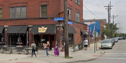 A street corner featuring a brick building with shops, pedestrians walking, and a mural on the adjacent wall.