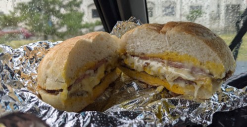 A close-up of a sandwich with layers of ham, cheese, and mayonnaise, wrapped in foil, sitting in a car. Rain is visible outside.