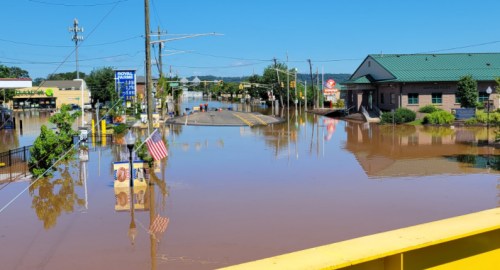 A flooded street with submerged buildings and signs, reflecting blue sky and power lines in the water.