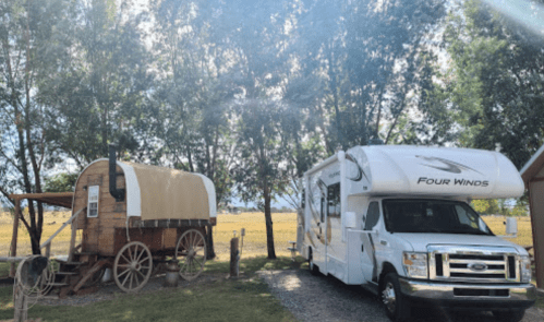 A vintage wagon and a modern RV parked side by side in a grassy area with trees and fields in the background.