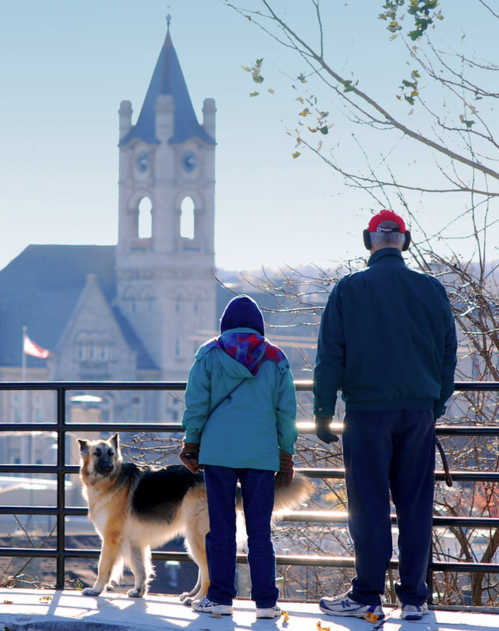 A person and a child stand by a railing with a dog, overlooking a historic building in the background on a clear day.