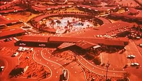 Aerial view of a circular hotel complex with winding roads and a central pool area, surrounded by palm trees.
