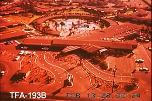 Aerial view of a circular building complex with a pool and surrounding roads, featuring palm trees and parked cars.