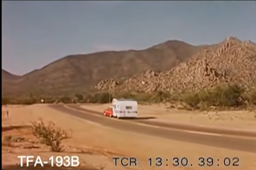 A white RV drives along a winding road with mountains and rocky terrain in the background under a clear blue sky.