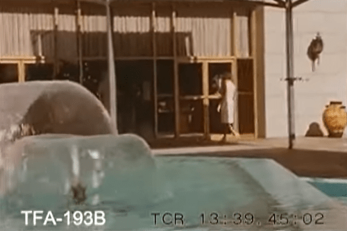A woman in a white dress walks past a fountain near a building entrance.