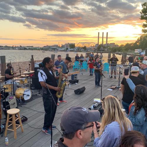 A lively outdoor concert at sunset, featuring musicians playing instruments with an audience enjoying the performance by the water.