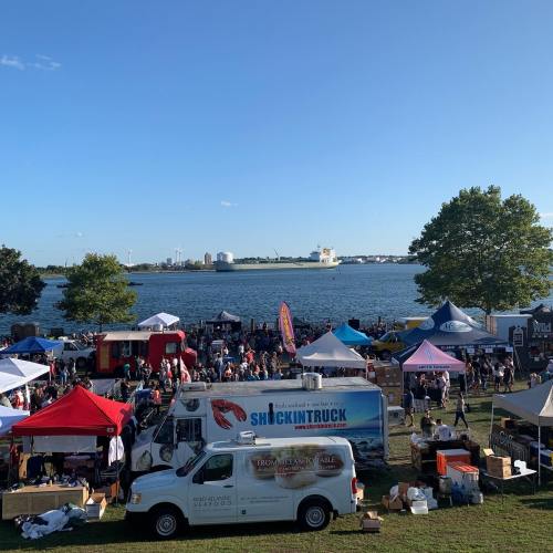 A vibrant outdoor market by the water, featuring food trucks, tents, and a crowd enjoying the sunny day.