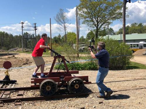A man in a red shirt rides a rail cart while another man takes photos nearby on a sunny day.
