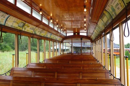 Interior of a vintage wooden tram, featuring rows of wooden benches and overhead lights, with a view of greenery outside.