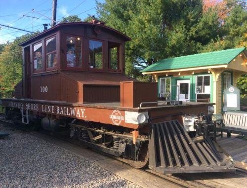 A vintage train engine with a wooden cabin, labeled "Shore Line Railway," parked near a green-roofed station.